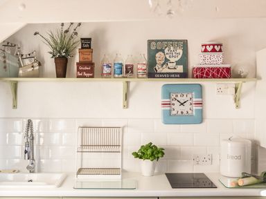 A kitchen counter with a sink a drying rack a plant an electric stove a bread container and a shelf with spice containers bottles a clock and decorative boxes at The Snug in Rescorla near St Austell