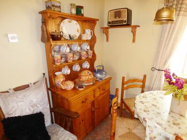 A dining area with wooden chairs a wooden cabinet with cups plates and teapots a table with floral tablecloth and flowers at Cookies Cottage Ballyshannon County Donegal