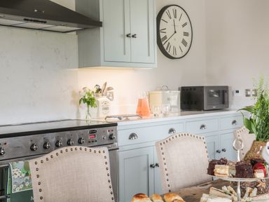 A kitchen with a stove oven and light blue cabinets with a clock on the wall and a dining table with chairs and food at Crane Field Laithe Otterburn North Yorkshire