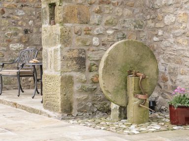 An outdoor area with a stone wall a large millstone a metal chair with a cushion and a small red flower pot at Crane Field Laithe Otterburn North Yorkshire