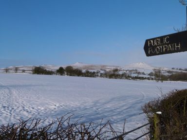 A snow-covered field with trees and hills under a clear blue sky with a public footpath sign in the foreground at Crane Field Laithe in Otterburn North Yorkshire