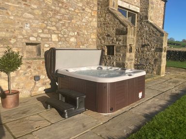 An outdoor hot tub with steps next to a stone house wall and a potted plant at Crane Field Laithe in Otterburn North Yorkshire