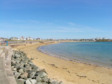 A sandy beach with a stone seawall and houses in the background at Anneddle in Four Mile Bridge