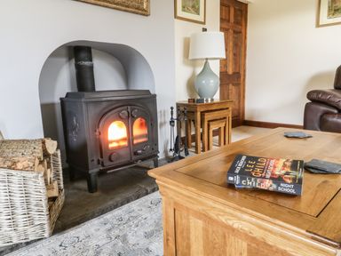 A wood burning stove with fire next to a basket of logs and a wooden table with a book and coasters at Hill Bank in Hutton Roof