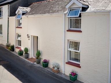 The front exterior of a cream house with six windows and six flower pots along the sidewalk at 3 Horse Pool Road in Laugharne