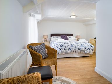 A bedroom with a bed with floral bedding two bedside tables with lamps a wicker chair with a cushion and a stool at 3 Horse Pool Road in Laugharne
