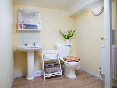 A bathroom with a pedestal sink mirror and soap dispensers next to a toilet with a wooden seat and a small shelf with towels and a basket at 3 Horse Pool Road in Laugharne