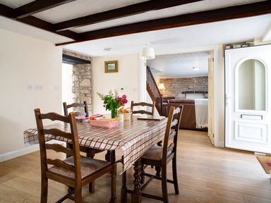 A dining area with a table covered by a checkered tablecloth and wooden chairs near an open door and a living room with brown sofas at 3 Horse Pool Road in Laugharne