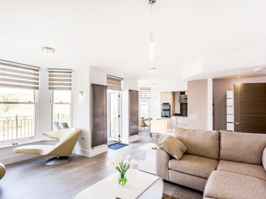 A living room with a sectional sofa coffee table with flowers a chair and kitchen in the background at No 6 Arlington Villas in Ilfracombe