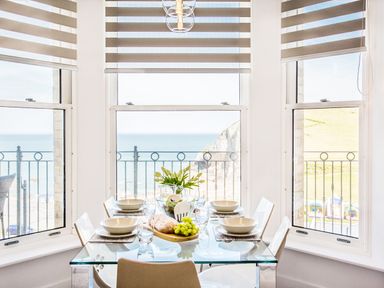 A dining area with a glass table set for four with a loaf of bread grapes and flowers by windows overlooking the sea at No 6 Arlington Villas in Ilfracombe