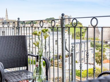A balcony with a wicker chair and flowers in a bottle overlooking buildings and a hillside at No 6 Arlington Villas in Ilfracombe