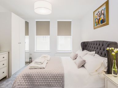 A bedroom with a gray upholstered bed with pillows and blankets two windows with blinds a dresser a wardrobe a nightstand with flowers and a painting on the wall at No 6 Arlington Villas in Ilfracombe
