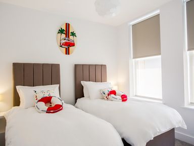 A bedroom with two single beds white bedding brown headboards surfboard wall art and windows with blinds at No 6 Arlington Villas Ilfracombe