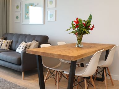 A dining area with a wooden table and white chairs next to a grey sofa with cushions at Little Fistral in Fistral