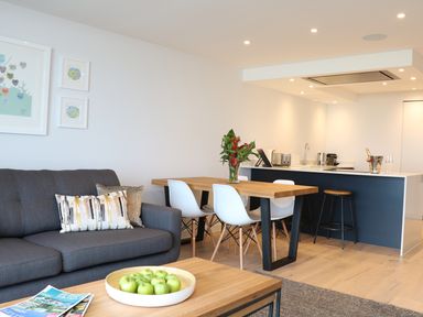 A living area with a gray sofa and patterned pillows a wooden dining table with white chairs and a kitchen island with stools at Little Fistral in Fistral
