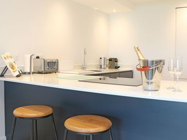 A kitchen counter with two wooden stools a toaster a coffee machine a bottle in a metal ice bucket and two glasses at Little Fistral in Fistral