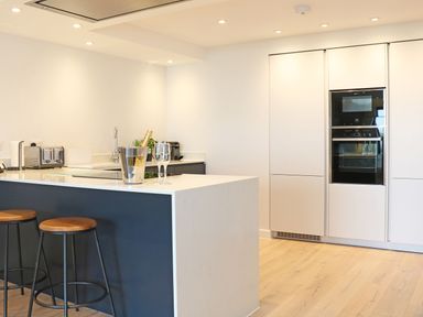 A kitchen with a white island counter black stools built in ovens and light wood flooring at Little Fistral in Fistral