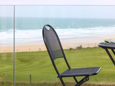 A black metal chair and a round table with sunglasses on a balcony overlooking the beach and ocean at Little Fistral in Fistral