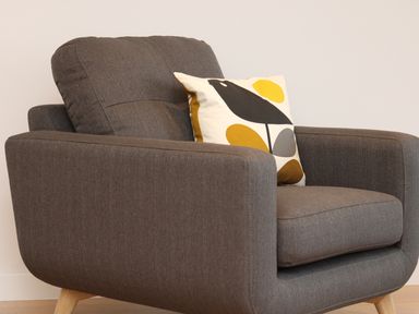 A grey fabric armchair with wooden legs and a decorative pillow featuring a black bird and colorful shapes at Little Fistral in Fistral