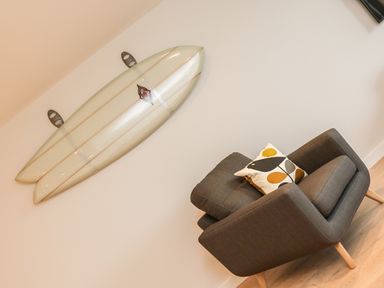 A surfboard mounted on a wall above a gray armchair with a patterned cushion at Little Fistral in Fistral