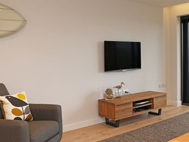 A living room with a grey armchair with a bird-patterned cushion a wall-mounted TV and a wooden TV stand at Little Fistral in Fistral