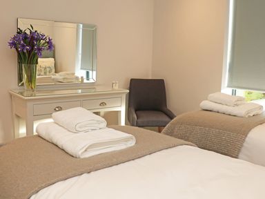 A bedroom with two beds covered with beige blankets and white towels on top a white dressing table with a mirror and a vase of purple flowers and a gray chair at Little Fistral in Fistral