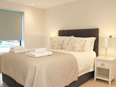 A bedroom with a bed covered with beige quilts and decorative pillows next to a nightstand with a lamp at Little Fistral in Fistral