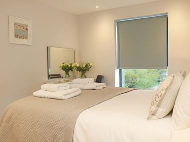 A bedroom with a bed, folded towels, a window with a blind, a mirror, and flowers on a dresser at Little Fistral in Fistral