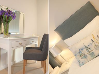 A white dressing table with a mirror and purple flowers in a vase next to a grey chair and a bedside table with a lamp and bed with a sailboat pillow at Little Fistral in Fistral