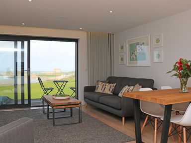 A living room with a dark sofa cushions wooden tables white chairs and sliding glass doors overlooking a lawn at Little Fistral in Fistral