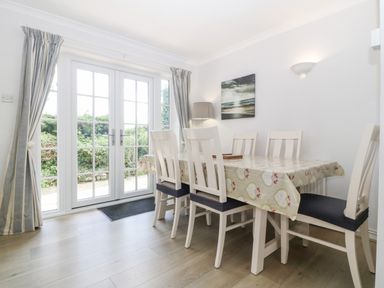 A dining room with a table covered by a patterned tablecloth and white chairs near glass doors and curtains at Glan Dwr in Red Wharf Bay