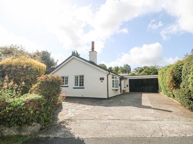 A single story house with a driveway and black garage doors surrounded by hedges and bushes at Glan Dwr in Red Wharf Bay