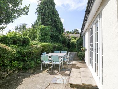 An outdoor patio area with a table and six chairs next to a white house with glass doors and surrounded by greenery at Glan Dwr in Red Wharf Bay
