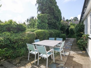 An outdoor patio area with a square table and six chairs next to a hedge and pathway at Glan Dwr in Red Wharf Bay