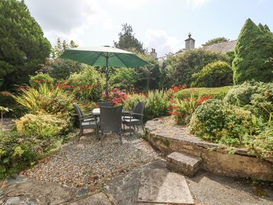 A garden area with a table and chairs under a green umbrella surrounded by bushes and flowers at Glan Dwr in Red Wharf Bay
