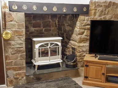 A stone fireplace with a white electric stove inside a living room with a wooden TV cabinet and a flat screen TV at Easterly Cottage in Bude