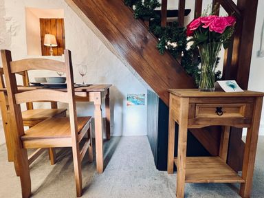 A dining area with a wooden table and chairs a small wooden side table with flowers under a wooden staircase at Easterly Cottage in Bude