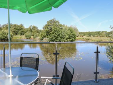 A patio table with chairs and a green umbrella overlooking a pond with trees and a field in the background at Lakeside Lodge in East Harling near Thetford