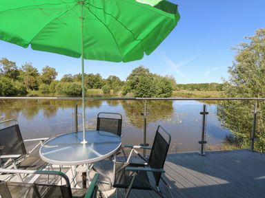 A glass table with green umbrella and black chairs on a deck overlooking a lake with trees at Lakeside Lodge in East Harling near Thetford