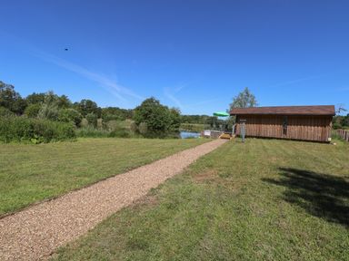 A gravel path leading to a wooden cabin near a pond with trees and a green umbrella at Lakeside Lodge in East Harling near Thetford