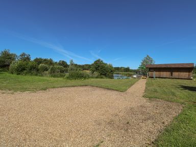 A gravel path leading to a wooden building near a pond with trees and grass at Lakeside Lodge in East Harling near Thetford