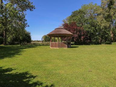A wooden gazebo with a thatched roof on a grass lawn surrounded by trees at Lakeside Lodge in East Harling near Thetford