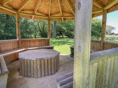 A wooden gazebo with built in benches and a wooden table in a garden at Lakeside Lodge in East Harling near Thetford