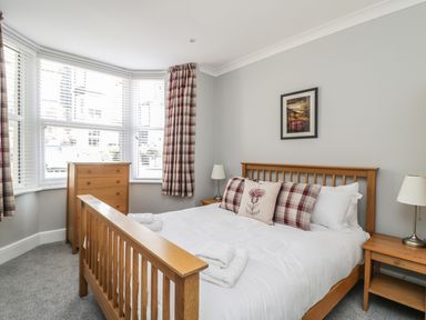 A bedroom with a wooden bed frame white bedding three pillows a chest of drawers nightstands with lamps and windows with plaid curtains at Ivy Bank in Windermere