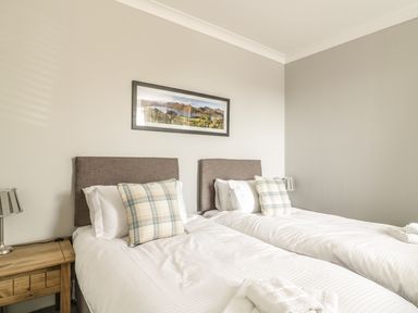 A bedroom with two single beds with plaid pillows a wooden bedside table with a lamp and a landscape painting on the wall at Ivy Bank in Windermere