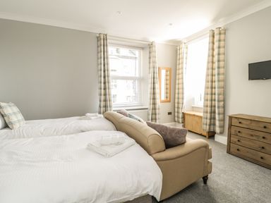 A bedroom with two single beds a beige sofa a wooden chest of drawers two windows with plaid curtains and a wall-mounted tv at Ivy Bank in Windermere