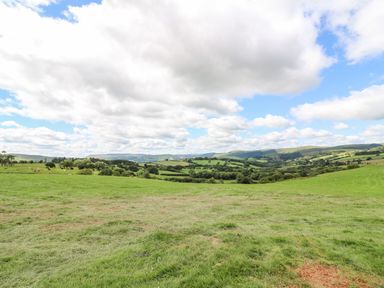 A grassy field with rolling hills and scattered trees under a partly cloudy sky at Meadow View in Rhayader