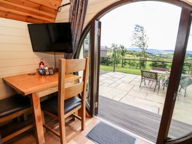 A wooden table and chairs next to a wall mounted television and an open curved door leading to an outdoor patio with a table and chairs at Meadow View in Rhayader