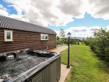 An outdoor area with a hot tub next to a wooden building and a grassy yard with lamp posts and outdoor furniture at Meadow View in Rhayader