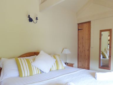 A bedroom with a wooden bed headboard with pillows and a bedside table with a lamp and a wooden door and a wall mirror at Old Winery Cottage in Golant near Fowey